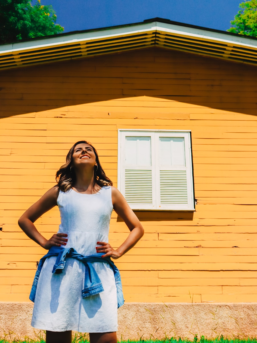 woman in white tank top and blue skirt standing in front of yellow house with homeowners insurance